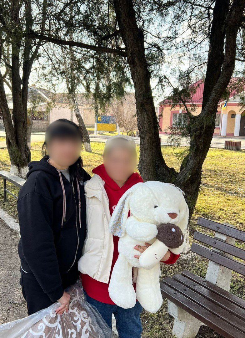 Двоє людей стоять на вулиці біля паркової лави у сонячний день. Одна особа тримає велику білу м'яку іграшку у вигляді кролика та іграшковий гриб. На фоні видно дерева, лаву та будівлі. Обличчя людей на фото приховані.