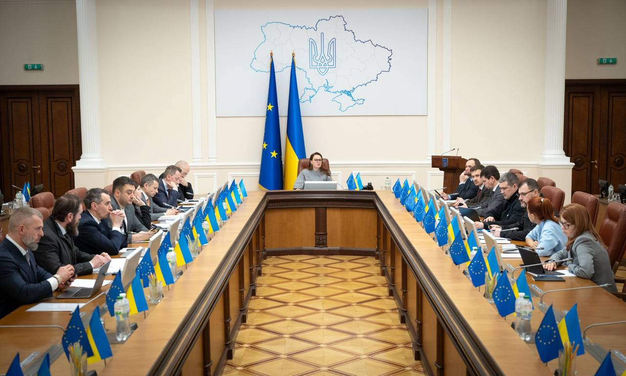 Робоча зустріч у залі засідань за великим П-подібним столом. Учасники сидять з обох боків, на столах встановлені прапорці України та Європейського Союзу. На центральній стіні розміщено карту України з гербом, перед якою розташоване місце головуючої особи.