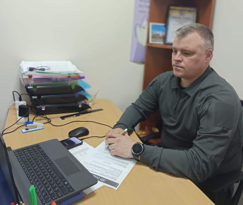 Чоловік у тактичній кофті сидить за робочим столом в офісі. Перед ним відкритий ноутбук, документи, комп'ютерна миша та смартфон. Він тримає ручку і дивиться вбік, на фоні видно полицю з папками та грамотами.