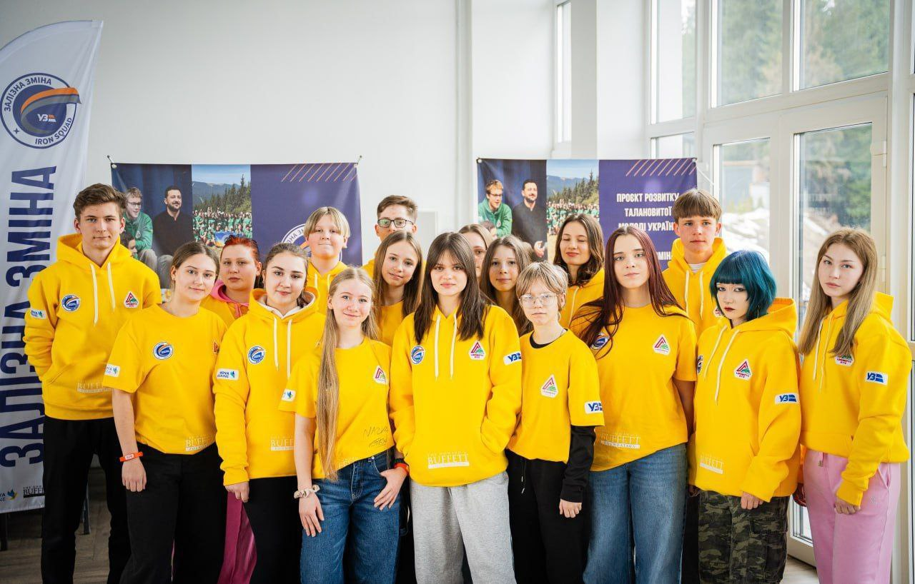 Група підлітків у яскраво-жовтому мерчі з логотипами стоїть у світлому приміщенні на фоні інформаційних банерів. Усі вони дивляться в камеру, позуючи для групового фото.