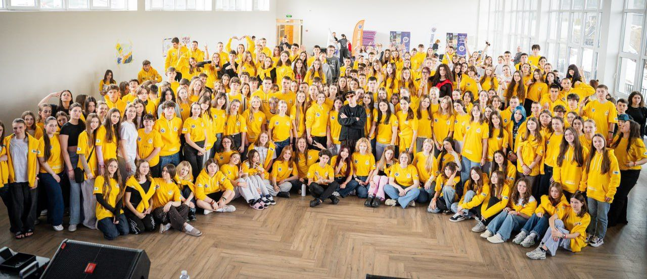 Групове фото великої кількості людей у просторому світлому залі. Більшість учасників одягнені в однакові яскраво-жовті футболки або худі. Люди стоять та сидять на підлозі, позуючи для спільного знімка.