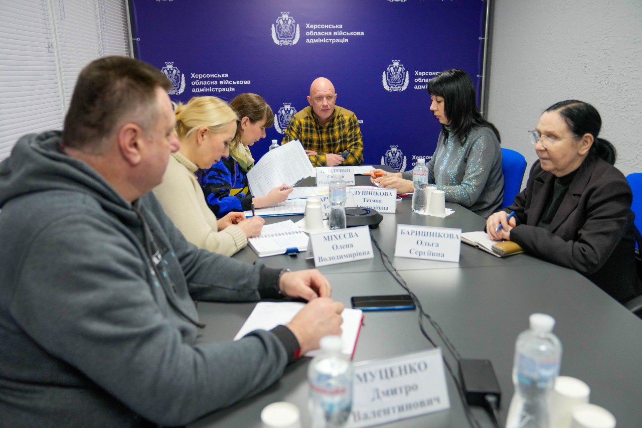 Група людей сидить за великим столом під час офіційної зустрічі або наради. Учасники працюють з документами на фоні синього брендованого банера Херсонської обласної військової адміністрації. На столі розміщені таблички з іменами та пляшки з водою.