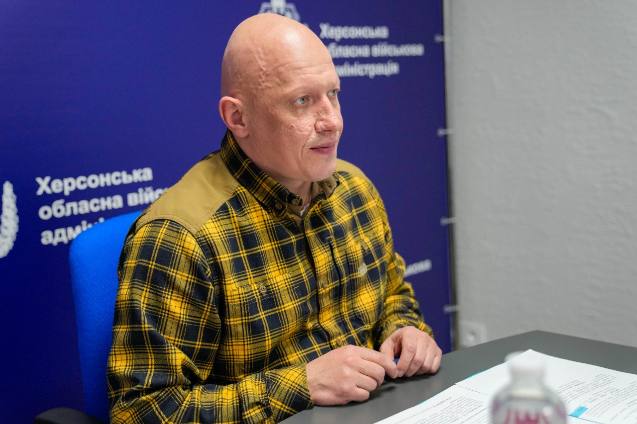 Чоловік у жовтій картатій сорочці сидить за столом на фоні синього банера з написами. Він дивиться вбік, склавши руки перед собою, а перед ним лежать документи.