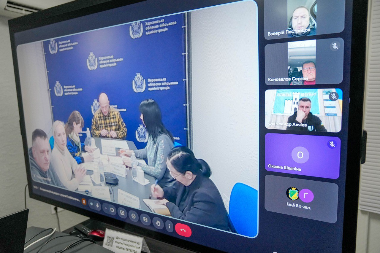 Скриншот онлайн-наради на великому моніторі. У головному вікні відображено групу людей за столом на фоні брендованого банера, а праворуч — стовпець із віконцями інших дистанційних учасників та іконками мікрофонів.