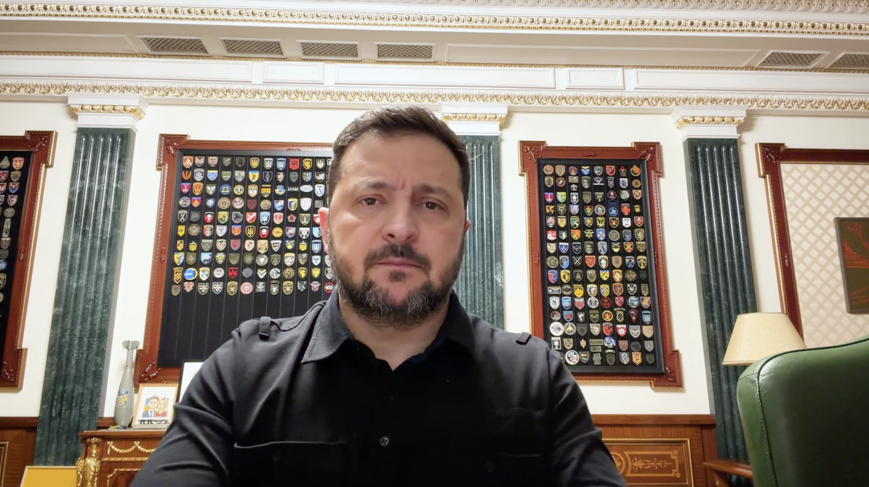 Чоловік у чорній тактичній сорочці сидить у кабінеті з класичним інтер'єром та колонами. На стіні за ним у рамках розміщені великі колекції військових шевронів. Він серйозно дивиться прямо в камеру, позуючи для відеозвернення.