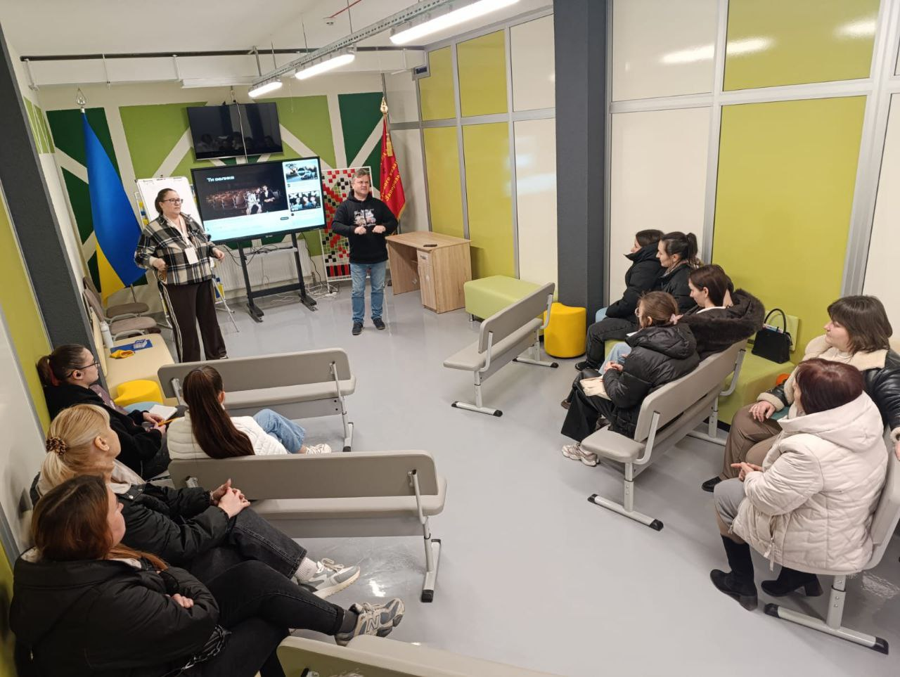 Група людей у сучасному світлому приміщенні під час презентації чи тренінгу. Двоє спікерів стоять біля великого екрана та державного прапора України, а слухачі розмістилися на лавах і стільцях навпроти них.