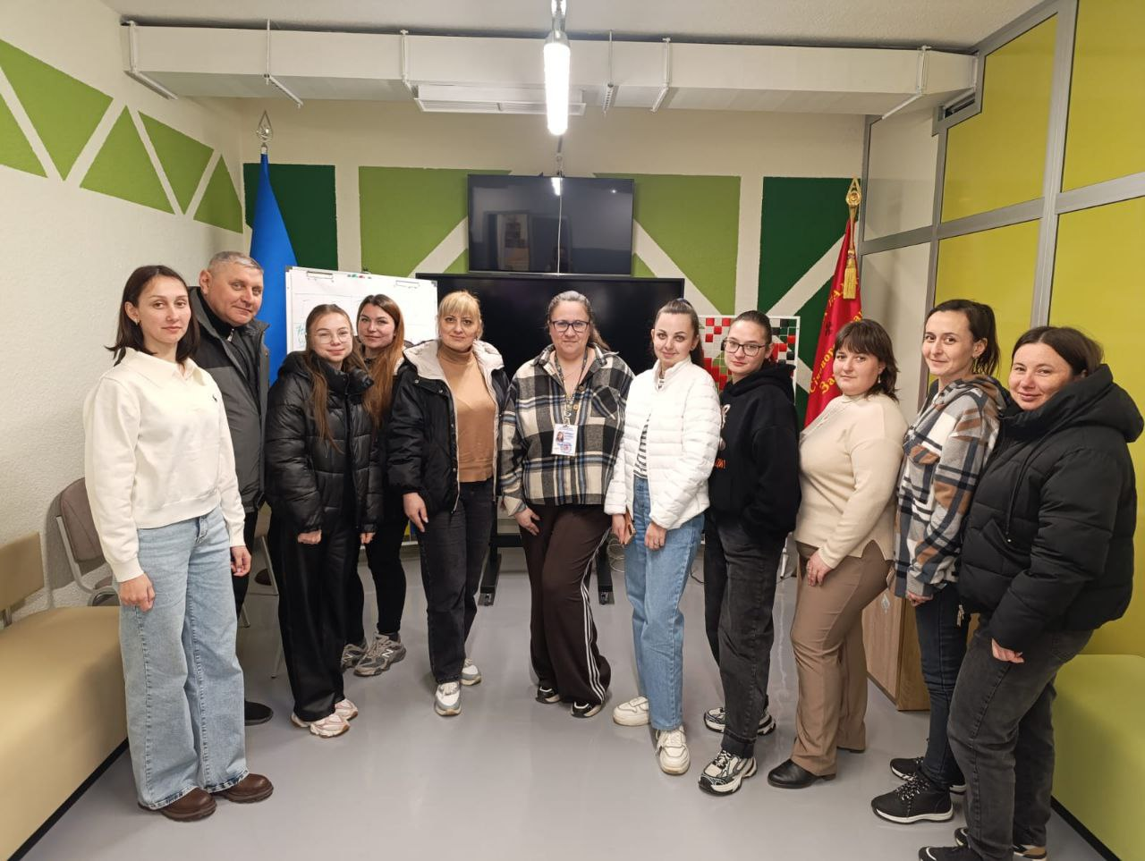 Група людей позирует для спільного фото в сучасному офісному або освітньому приміщенні. Вони стоять на фоні великого екрана, білої дошки та державного прапора України, створюючи атмосферу завершення робочої зустрічі чи тренінгу.