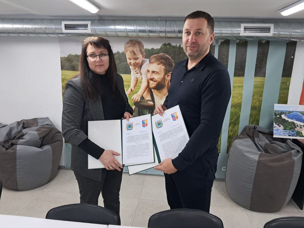 Жінка та чоловік у сучасному приміщенні тримають розгорнуті офіційні документи з печатками та гербами. Вони стоять на фоні стіни з великим фотопринтом щасливої родини та м'яких крісел-мішків.