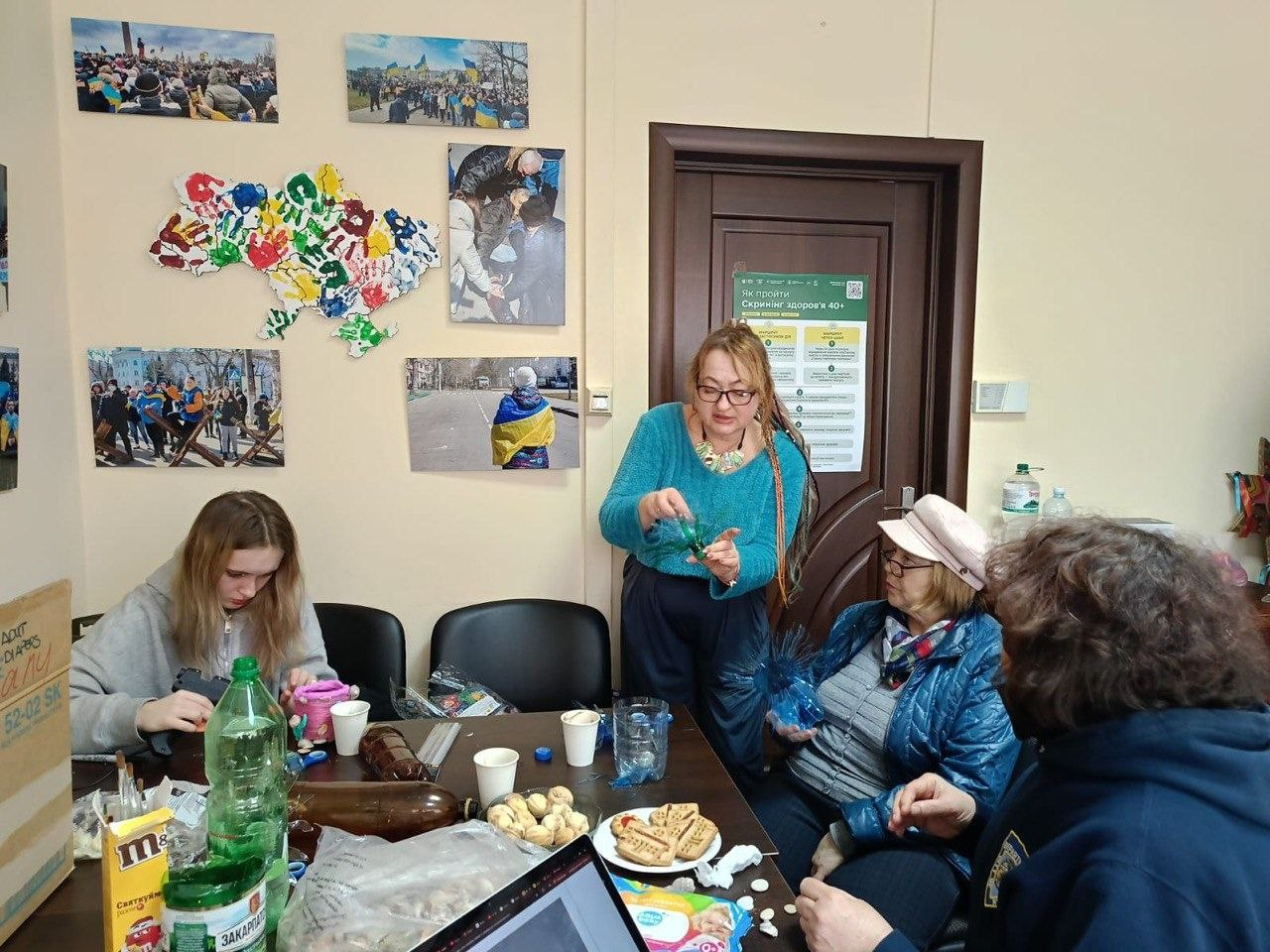 Люди за столом у волонтерському штабі займаються творчістю на фоні патріотичних плакатів. У центрі жінка демонструє виріб зі стрічок; на столі лежать підручні матеріали та солодощі.