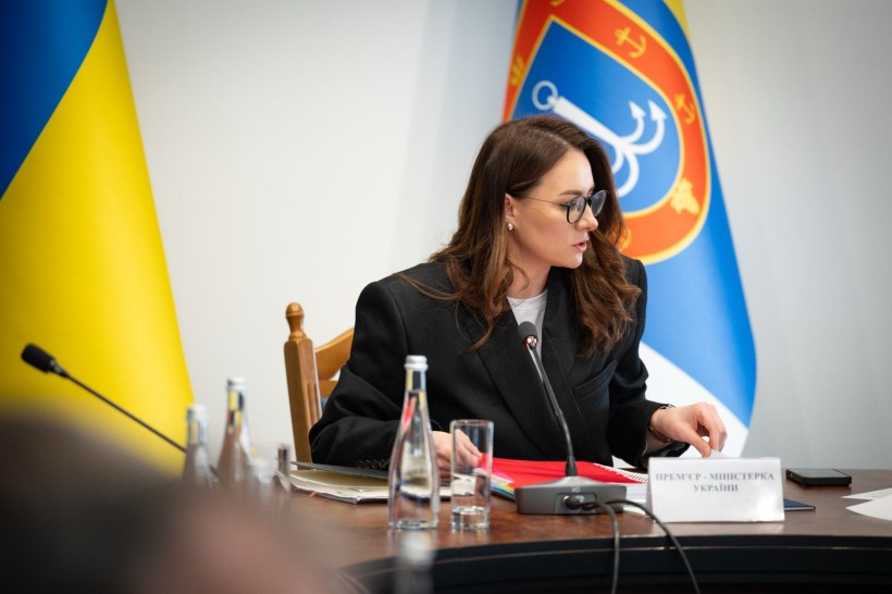 Жінка в окулярах та офіційному чорному піджаку сидить за робочим столом під час наради. Перед нею лежать папери, стоїть мікрофон та скляна пляшка води. На фоні видно державний прапор України та синій стяг із гербом.