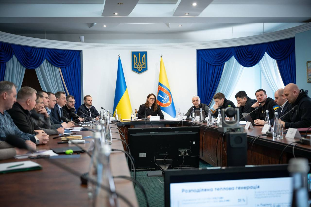 Офіційна нарада за великим підковоподібним столом під головуванням жінки. На фоні — прапор України та стяг із гербом, на столах — мікрофони, ноутбуки та документи.