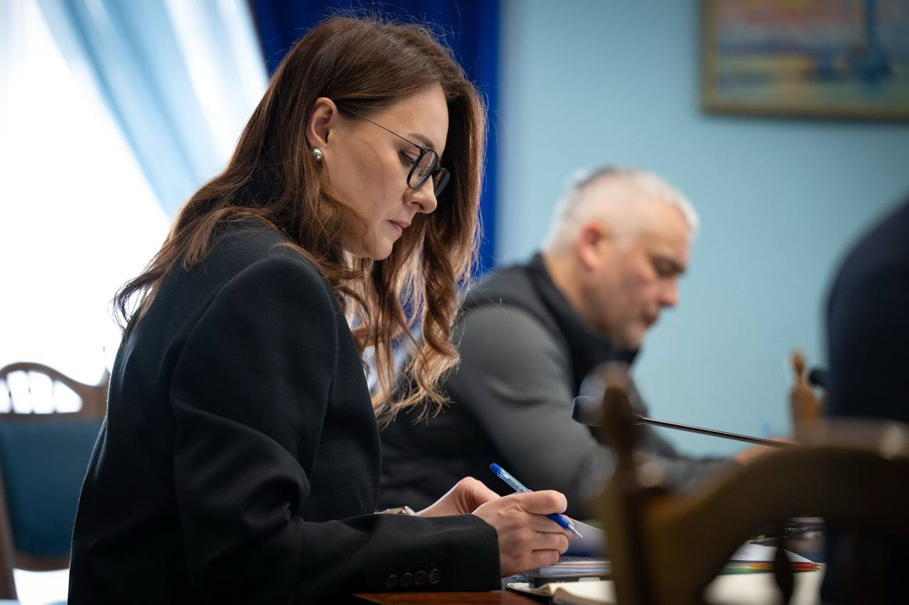 Жінка в окулярах та чорному піджаку зосереджено робить записи ручкою під час ділової зустрічі. На фоні, з невеликим розмиттям, видно чоловіка та мікрофони на столі, що вказує на офіційну обстановку наради.