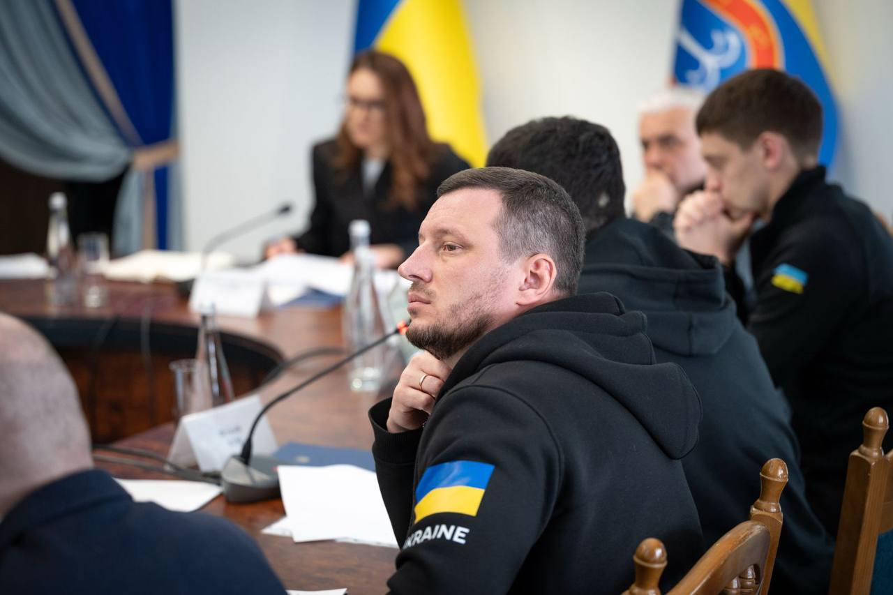 Група людей у темному одязі сидить за круглим столом під час офіційної наради. Чоловік на передньому плані має нашивку з прапором України на рукаві. На фоні видно жінку в окулярах, державну символіку та мікрофони.