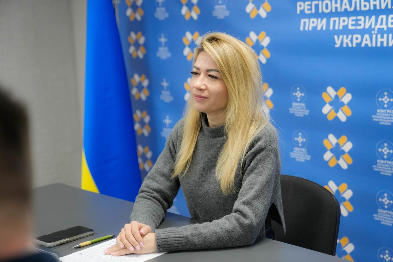 Жінка за столом під час засідання на фоні банера Конгресу місцевих та регіональних влад і прапора України.