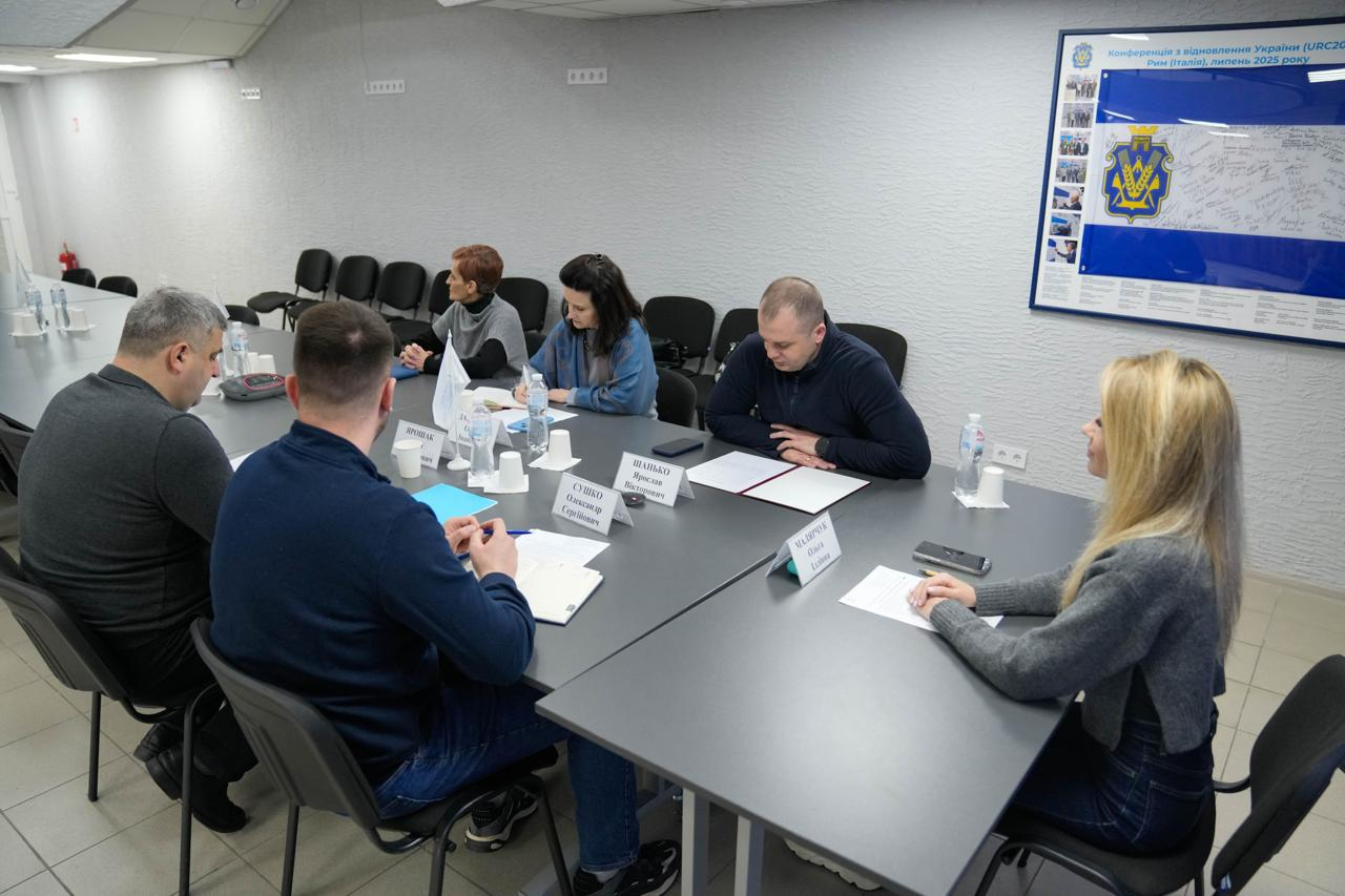 Група людей за робочим столом під час ділової зустрічі в офісному приміщенні. На стіні розміщено плакат, присвячений Конференції з відновлення України.