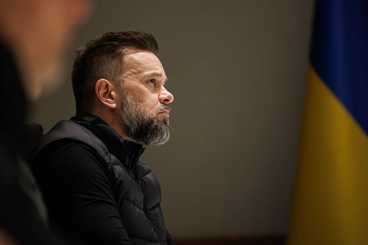 Чоловік із бородою у профілі на офіційному заході на фоні державного прапора України.