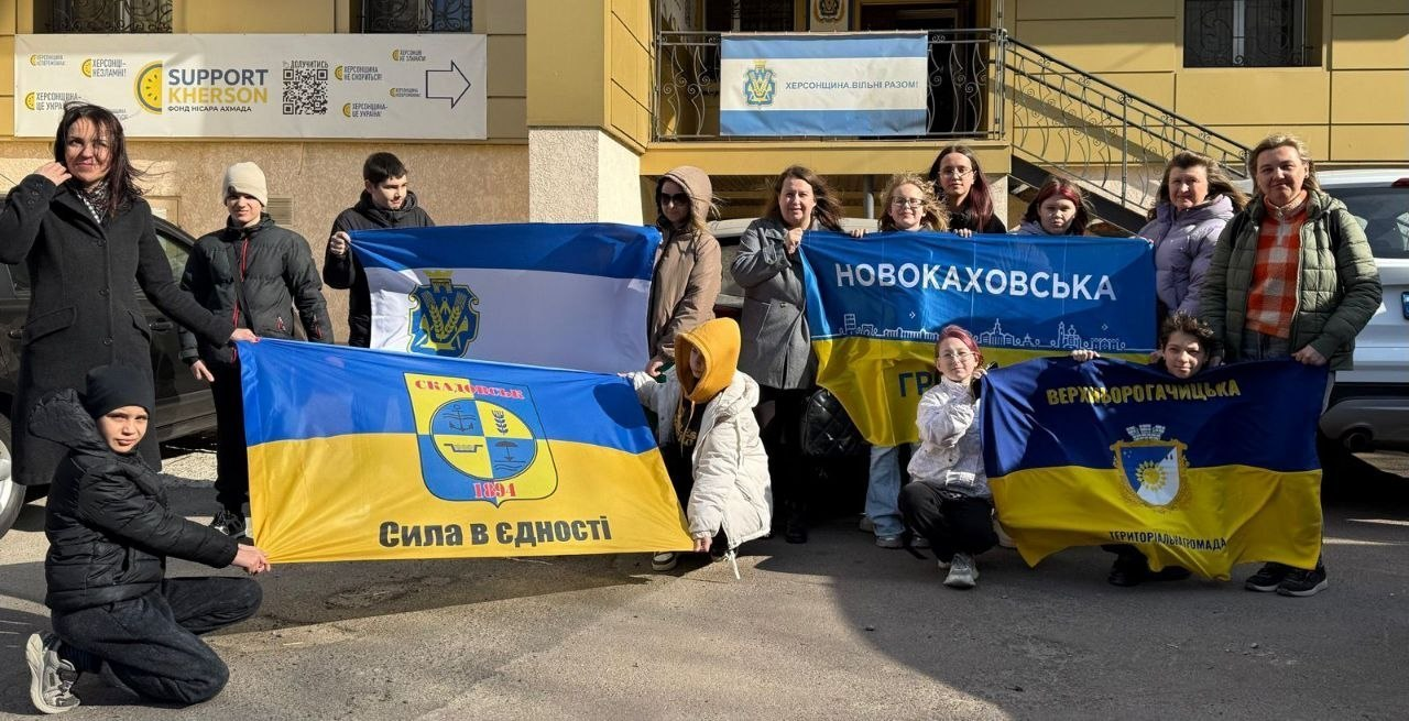 Група людей перед будівлею тримає прапори України та громад Херсонщини (Скадовської, Новокаховської, Верхньорогачицької). На фоні — плакати на підтримку Херсона.