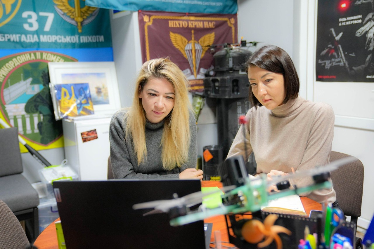 Дві жінки працюють за ноутбуком у приміщенні з військовою символікою та безпілотником на передньому плані. На стінах видно прапори 37-ї бригади морської піхоти та Десантно-штурмових військ.