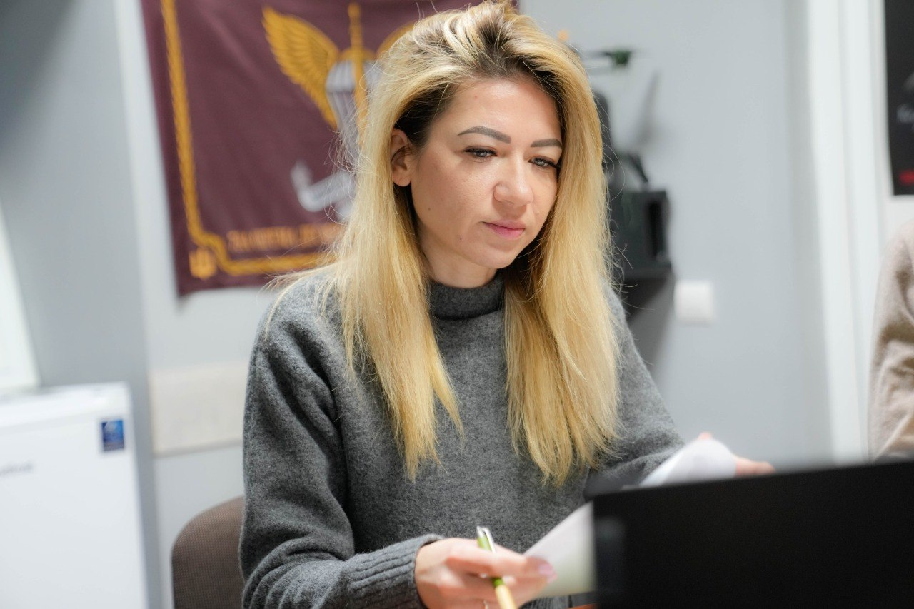 Жінка у сірому светрі працює з документами за робочим столом. На фоні видно частину прапора Десантно-штурмових військ України.