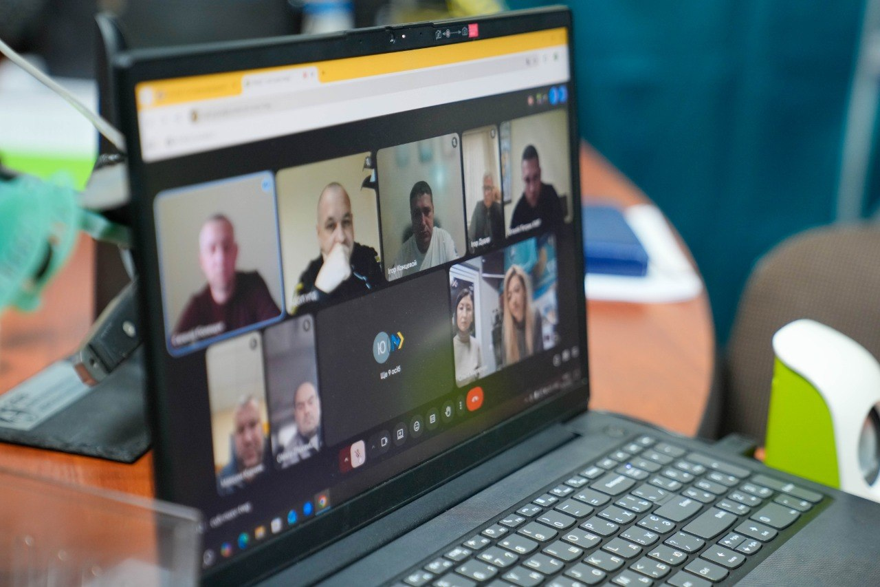 На екрані відкритого ноутбука відображається онлайн-нарада у форматі відеоконференції з багатьма учасниками. Пристрій стоїть на робочому столі в офісному приміщенні.