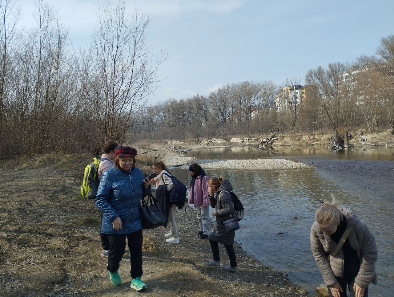 Група людей на прогулянці біля річки у прохолодну погоду. На задньому плані видно голі дерева та міські будівлі, а на передньому плані люди в демісезонному одязі відпочивають на кам'янистому березі біля води.