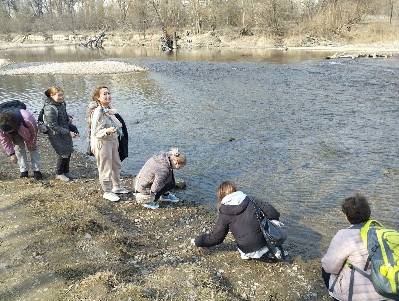 Група людей відпочиває на березі мілководної річки в оточенні весняного або осіннього лісу. Дехто розглядає воду або каміння біля краю, інші просто стоять поруч. Усі одягнені в теплий повсякденний одяг і куртки.
