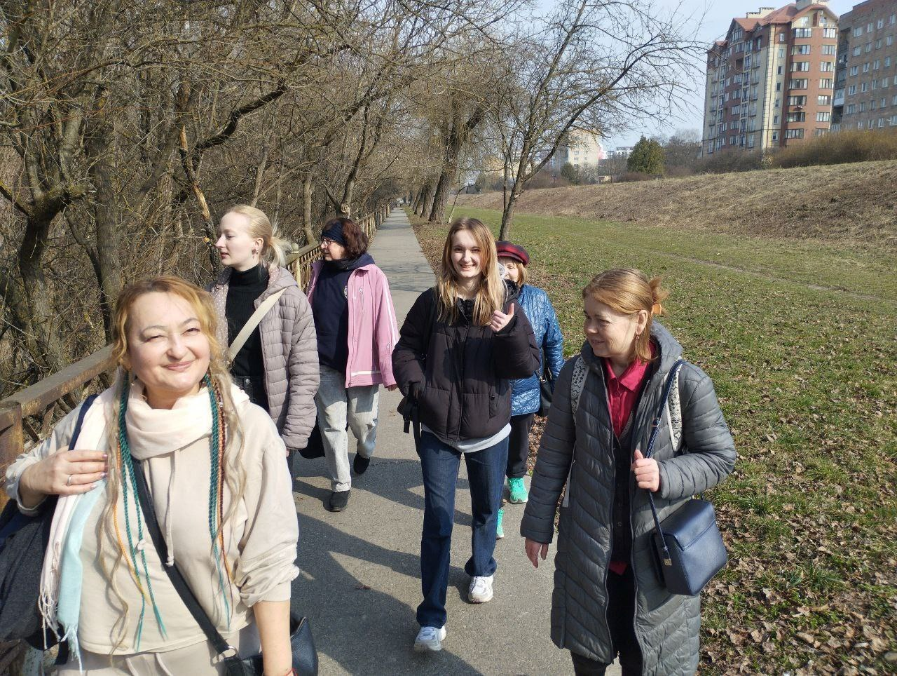 Група жінок різного віку прогулюється парковою доріжкою вздовж річки. Вони одягнені в демісезонні куртки та светри. Поруч видно схил із сухою травою, голі дерева та багатоповерхові будинки вдалині.