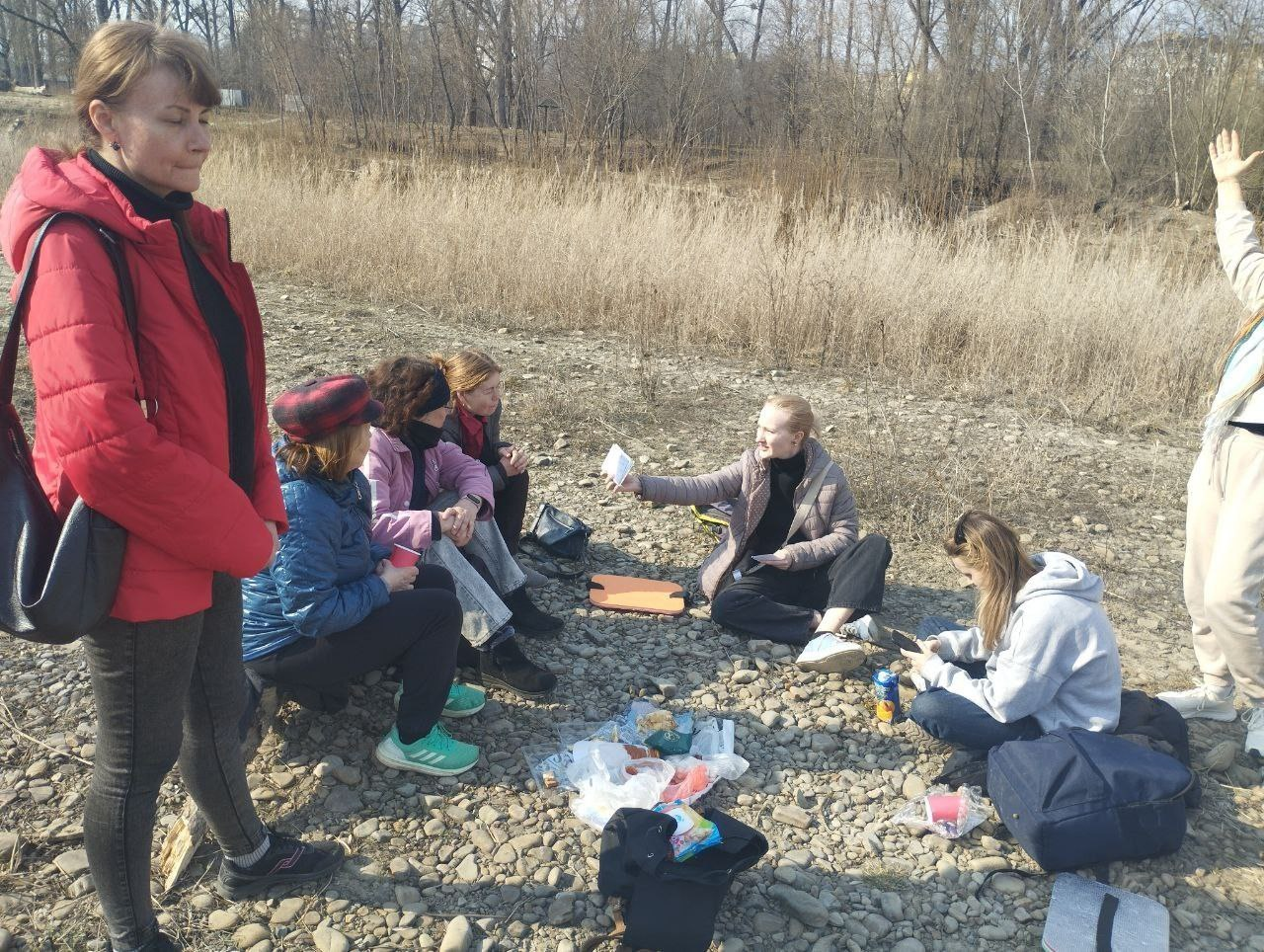 Група людей влаштувала пікнік на кам'янистому березі річки в прохолодну пору року. Вони сидять колом на землі поруч із їжею та речами, спілкуються та відпочивають. На фоні видно суху траву та безлисті дерева.