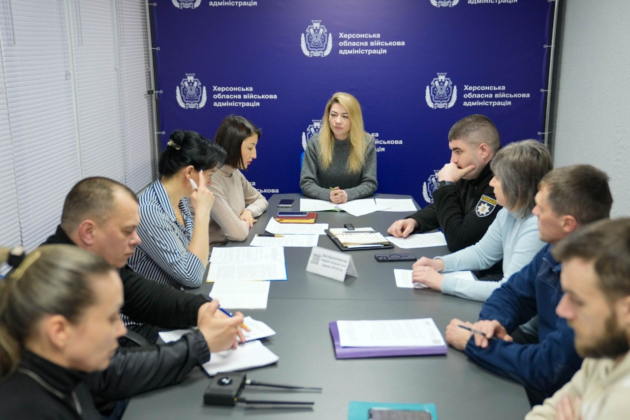 Група людей проводить офіційну робочу зустріч за великим столом. Учасники обговорюють документи, роблять записи в блокнотах та користуються смартфонами. Зустріч проходить у кабінеті на фоні синього банера з офіційною символікою.