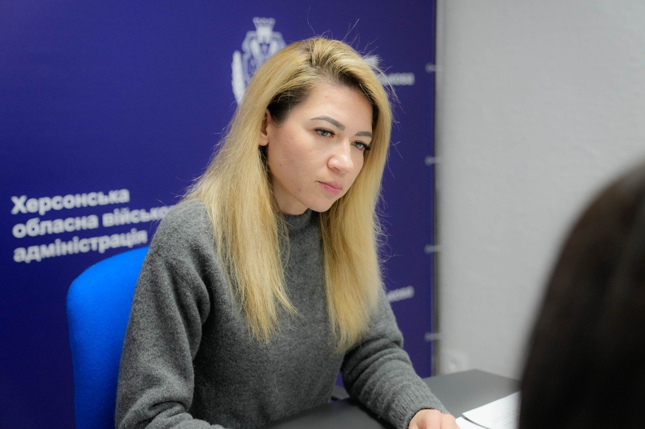 Жінка зі світлим волоссям у сірому светрі зосереджено слухає співрозмовника під час офіційної зустрічі. Вона сидить за столом на фоні синього банера з державною символікою та офіційним написом.