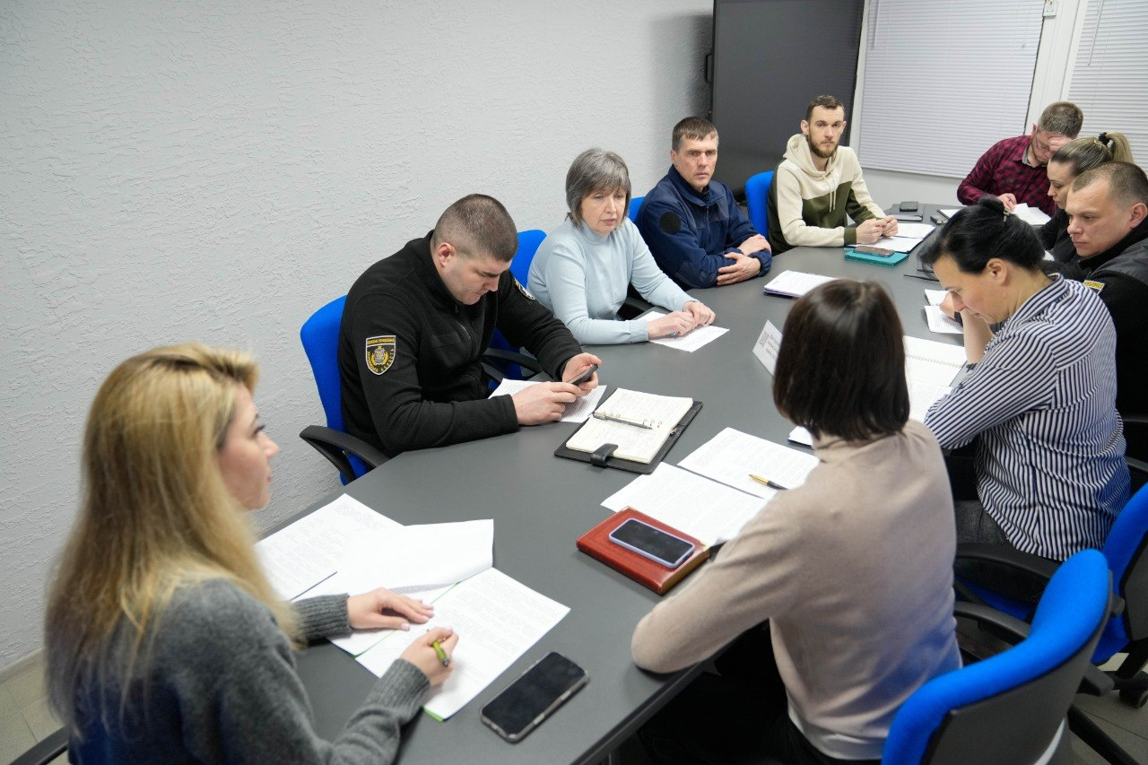 Група людей проводить робочу нараду в офісному приміщенні. Учасники сидять за довгим столом, на якому лежать документи, блокноти та смартфони. Серед присутніх є особи у цивільному одязі та у формі з шевронами.