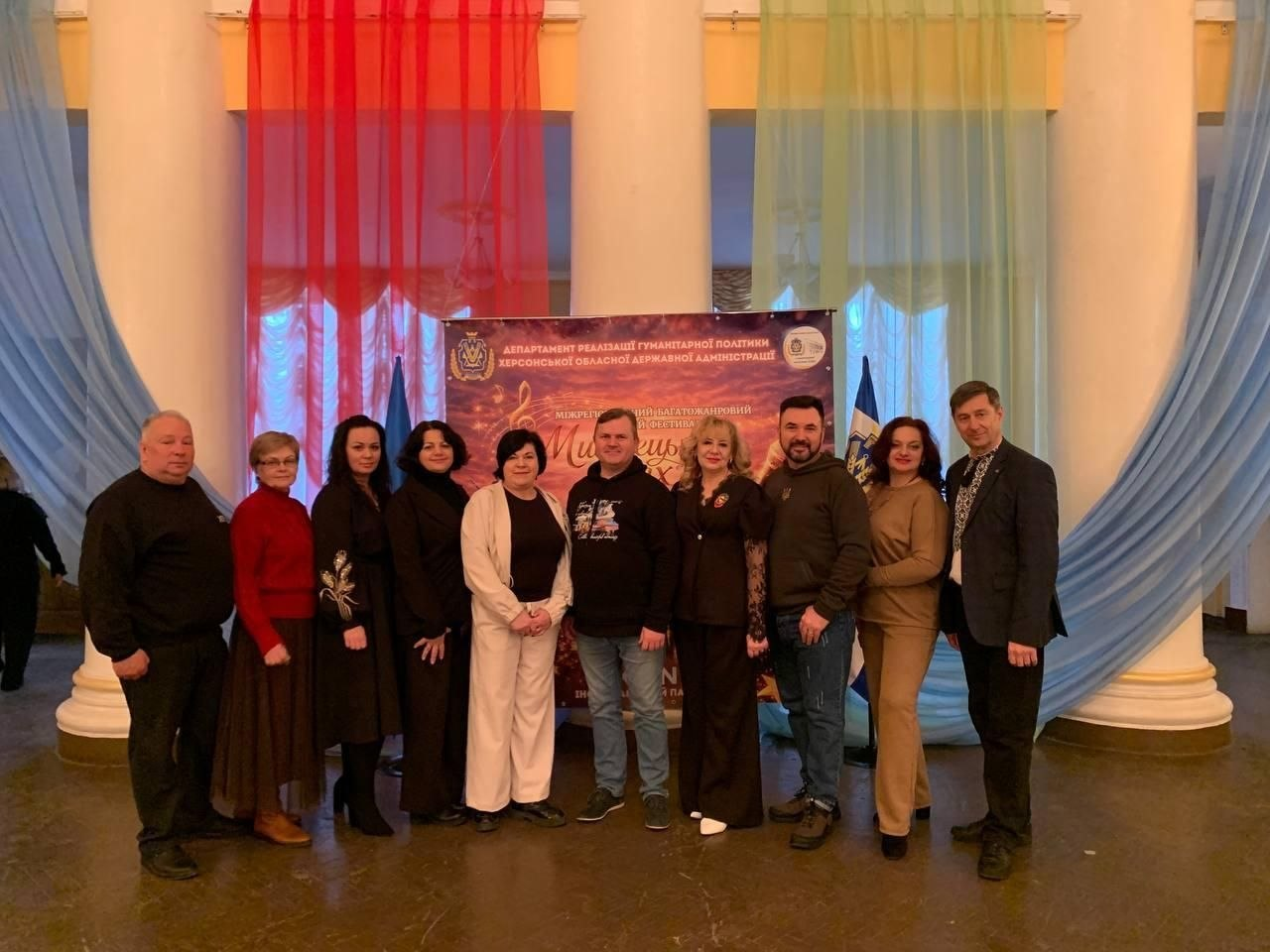 Група людей стоїть у ряд для спільного фото в урочистій залі. На задньому плані розташований великий інформаційний банер та декоративні штори червоного, блакитного та світло-зеленого кольорів. Учасники події одягнені в офіційно-діловий та повсякденний одяг.