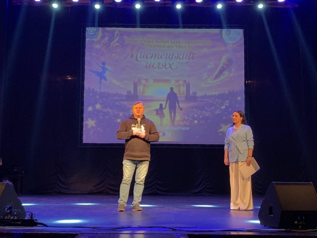 На освітленій сцені чоловік із мікрофоном та жінка з документами проводять захід. На задньому плані — великий екран із назвою творчого фестивалю «Мистецький шлях» та зображенням дитини з дорослим.