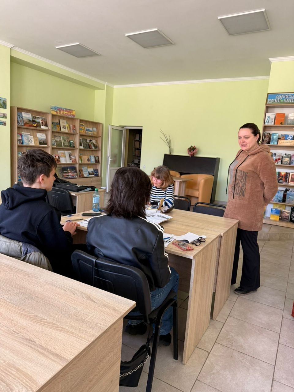 Група людей сидить за столами у світлій бібліотеці або навчальному класі. Жінка в коричневому худі стоїть поруч і спілкується з присутніми. На задньому плані видно стелажі з книгами та журналами.