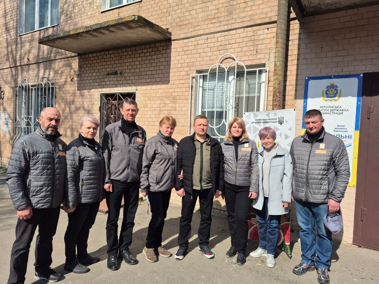 Група з восьми дорослих людей стоїть у ряд перед цегляною будівлею у сонячний день. Більшість із них одягнені в однакові сірі куртки. На стіні будівлі поруч із ними розміщено інформаційні плакати з державною символікою України.