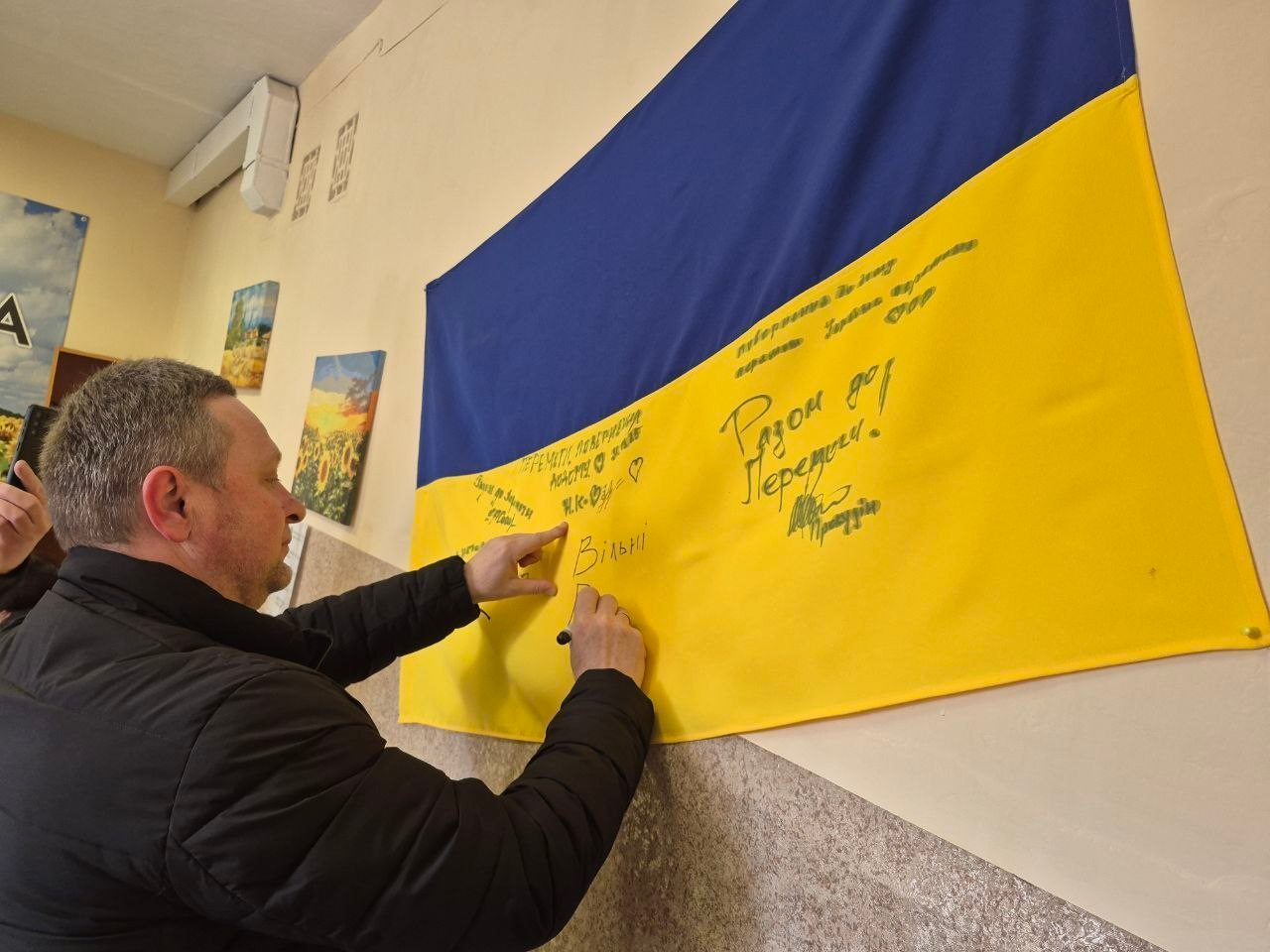 Чоловік у темній куртці пише побажання на великому державному прапорі України, закріпленому на стіні в приміщенні. На жовтій частині прапора вже видно кілька написів маркером, зокрема фразу «Разом до Перемоги!». На стіні поруч висять невеликі картини.