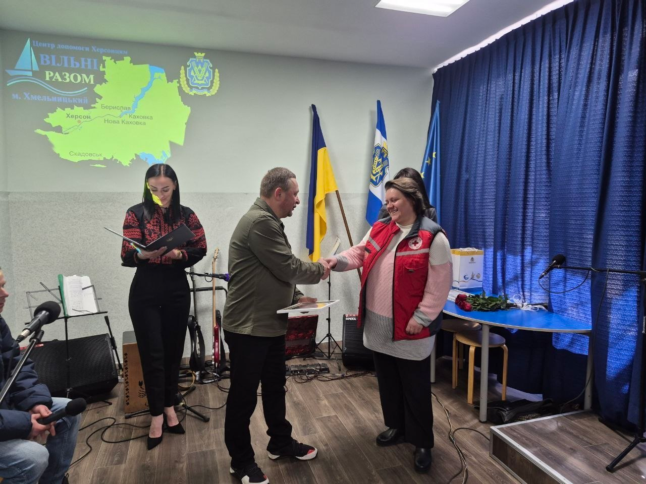 У приміщенні з прапорами та проєкцією карти Херсонщини на стіні чоловік вручає подяку або грамоту жінці в жилетці Червоного Хреста. Поруч стоїть жінка у вишиванці з папкою в руках. На задньому плані видно музичне обладнання та сині штори.
