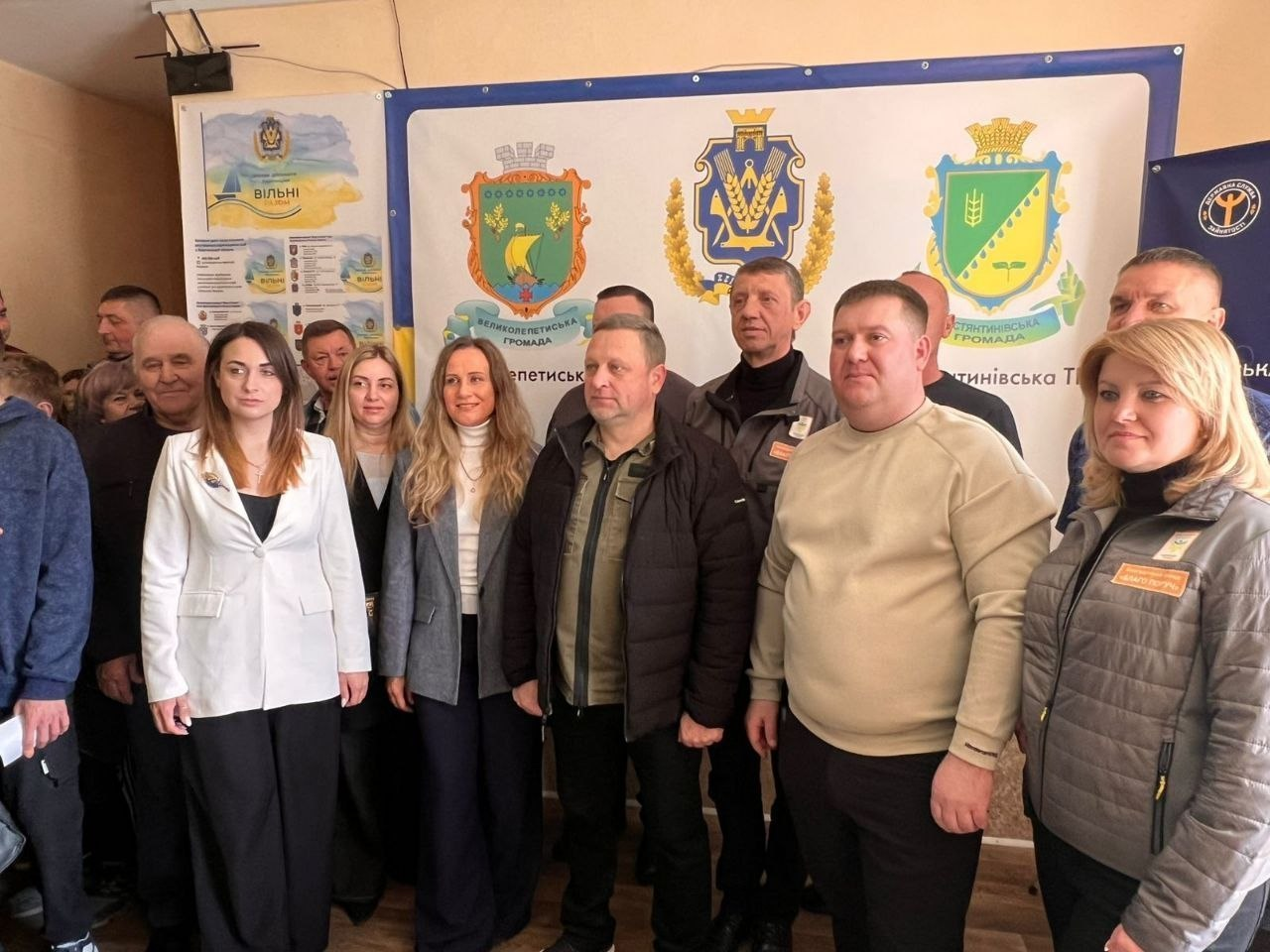 Група чоловіків та жінок стоїть у приміщенні на фоні великого банера з гербами територіальних громад. Деякі присутні одягнені в офіційний одяг, інші — у повсякденні куртки або світшоти. На задньому плані ліворуч також видно інформаційний плакат із державною символікою та написом «Вільні».