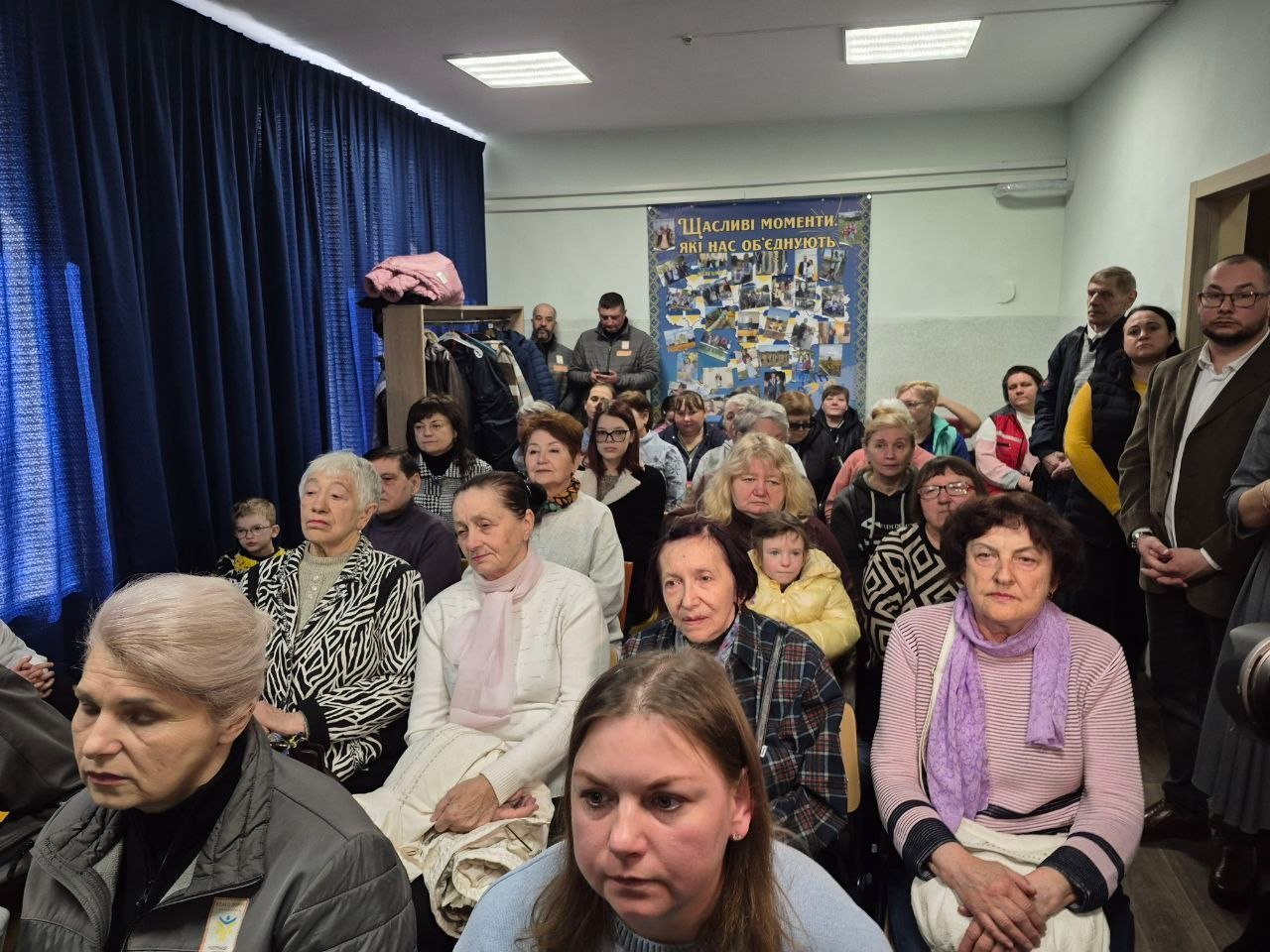 У приміщенні з синіми шторами зібралася велика група людей різного віку. На стіні за ними висить колаж із фотографіями під назвою «Щасливі моменти, які нас об'єднують». Люди сидять на стільцях та стоять у проходах, уважно спостерігаючи за подією поза кадром.