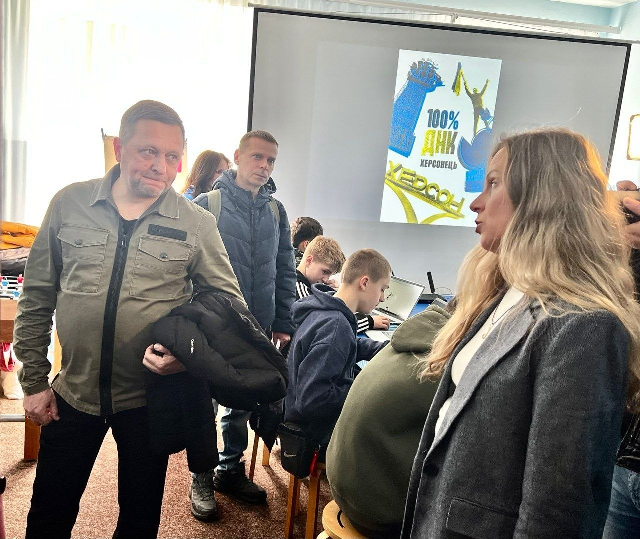 У навчальному центрі група дорослих та підлітків зібралася навколо проєкційного екрана. На екрані видно плакат із написом «100% ДНК Херсонець». Підлітки працюють за ноутбуками, а на передньому плані чоловік у куртці кольору хакі та жінка в сірому піджаку ведуть розмову.
