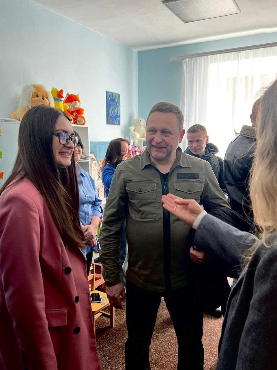 У світлій кімнаті з блакитними стінами група людей зібралася для спілкування. На передньому плані жінка в рожевому піджаку та окулярах посміхається чоловіку в куртці кольору хакі. На задньому плані видно полиці з м'якими іграшками, що вказує на дитячий простір або ігрову кімнату.