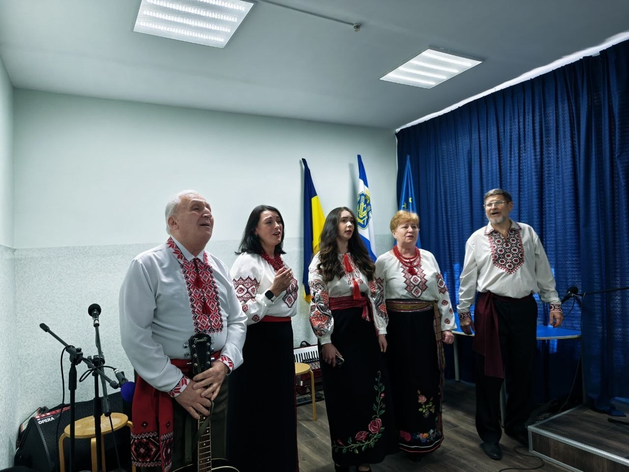 Ансамбль із п'яти виконавців у традиційному українському вбранні — вишиванках та шароварах — співає в приміщенні з синіми шторами. На передньому плані чоловік тримає гітару, а поруч із групою стоять мікрофони. На задньому плані на стіні встановлені державний прапор України та прапор Херсонської області.