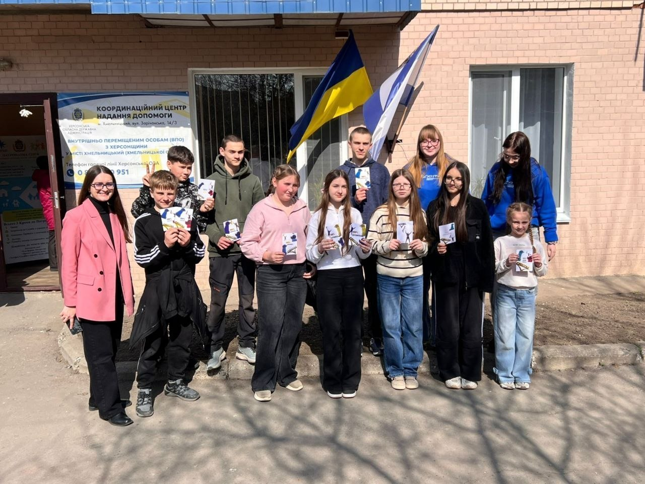 Група дітей та дорослих стоїть на вулиці перед будівлею Координаційного центру надання допомоги ВПО з Херсонщини. Вони тримають у руках невеликі книжки або брошури. Позаду них на стіні закріплені прапори України та Херсонської області, а поруч із входом розміщено інформаційний стенд із контактами гарячої лінії.