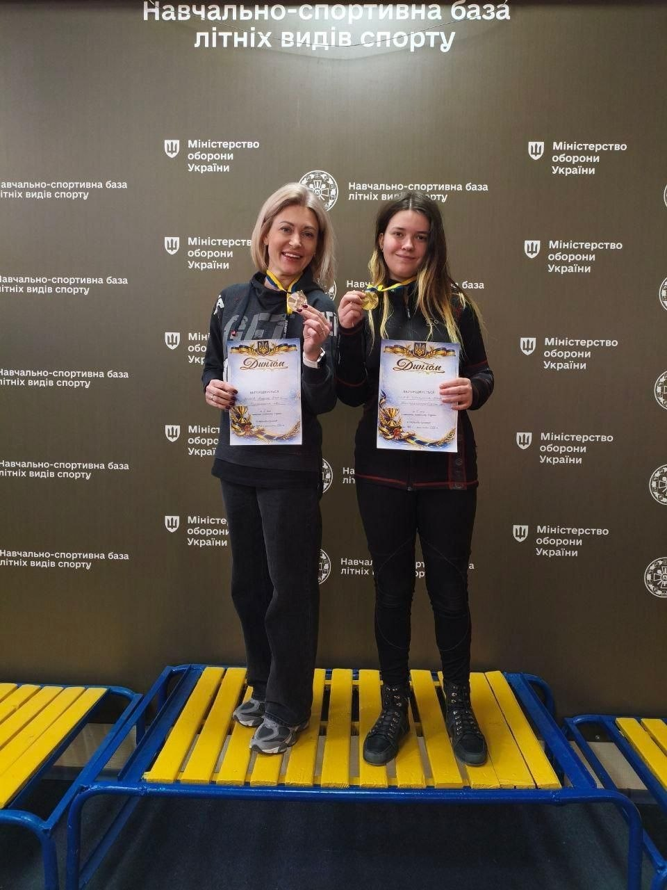Дві жінки стоять на синьо-жовтому спортивному п'єдесталі, тримаючи в руках нагородні дипломи та золоті медалі. Вони позують на фоні темного банера з логотипами Міністерства оборони України та повторюваним написом «Навчально-спортивна база літніх видів спорту».