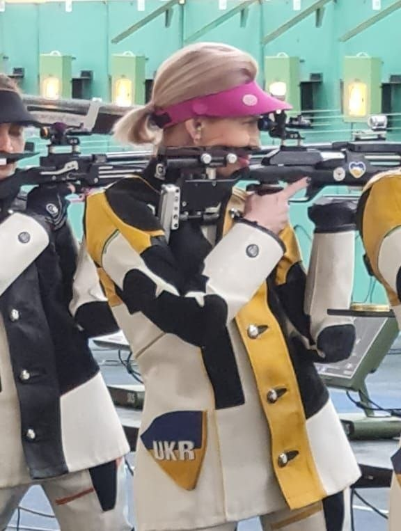 Спортсменка в професійній стрілецькій екіпіровці жовто-біло-чорного кольору з абревіатурою «UKR» на кишені цілиться з гвинтівки. На голові у неї яскраво-рожевий козирок. На задньому плані видно обладнане стрільбище з мішенями та іншими учасниками змагань.