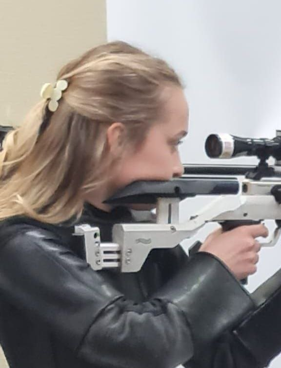 Спортсменка в чорній шкіряній куртці для стрільби цілиться з пневматичної гвинтівки марки Walther. Її обличчя щільно притиснуте до щоки приклада, а погляд спрямований через діоптричний приціл. Волосся дівчини зібране світлою шпилькою.