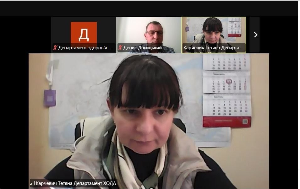 Скриншот вікна відеоконференції у форматі Zoom. На основній частині екрана зображена жінка, яка сидить в офісному приміщенні на фоні настінного календаря. У верхній частині вікна видно іконки інших учасників: одного чоловіка, ще одне вікно з тією ж жінкою та графічне зображення з літерою «Д» на червоному фоні.