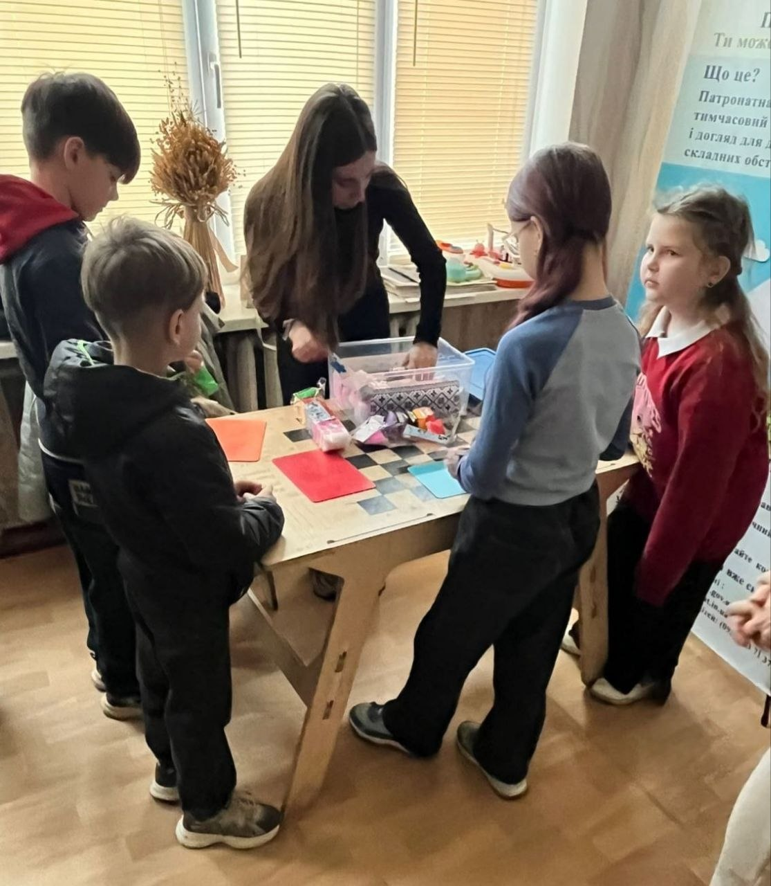 Група дітей зібралася навколо столу, за яким дівчина-підліток дістає приладдя для творчості з прозорого контейнера. На столі лежать кольорові аркуші паперу та канцелярські товари. На задньому плані видно вікно з жалюзі, дідух на підвіконні та інформаційний банер про патронатну допомогу.