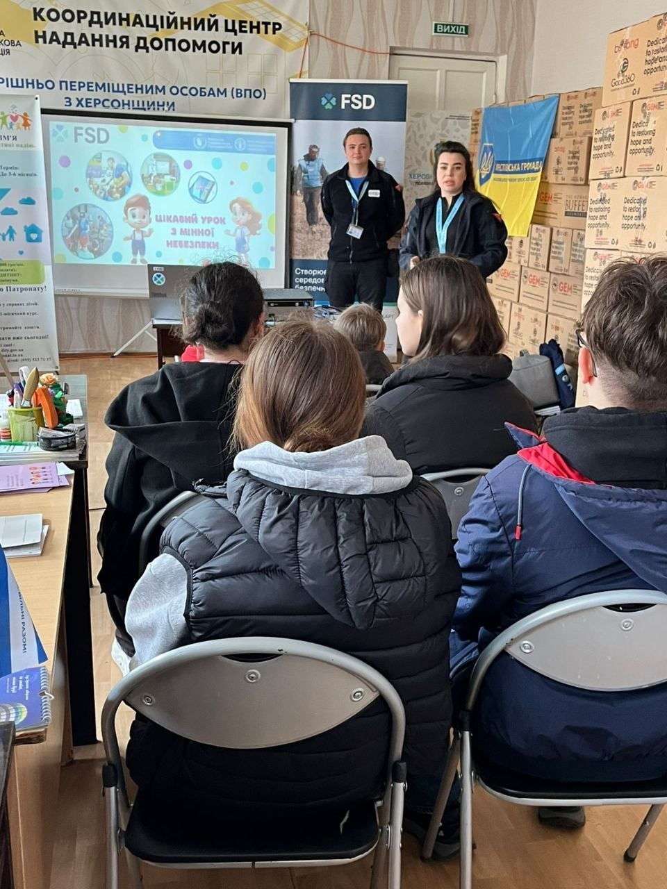 У приміщенні Координаційного центру допомоги ВПО з Херсонщини двоє лекторів у формі організації FSD проводять заняття для групи підлітків. На екрані проєктора видно заголовок: «Цікавий урок з мінної небезпеки». На стіні поруч висить український прапор, а праворуч складені коробки з гуманітарною допомогою.