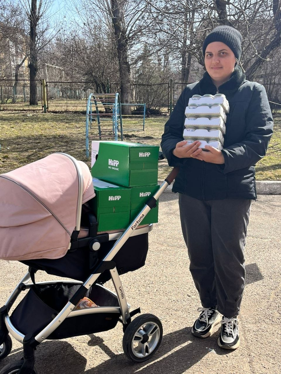 На вулиці жінка в темній куртці та шапці тримає в руках упаковку дитячого харчування. Поруч із нею стоїть рожева дитяча коляска, на якій складені зелені коробки з логотипом бренду HiPP. Подія відбувається на відкритому майданчику з деревами на фоні.